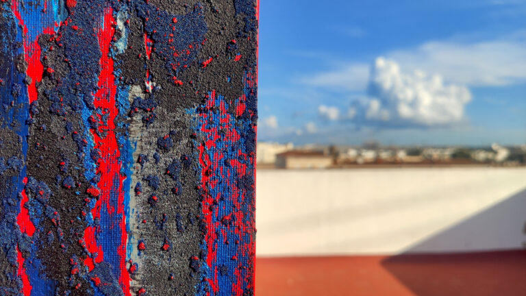 Close-up of a textured abstract painting with bold blue and red layers, rough granular surface, and high-contrast vertical strokes.
